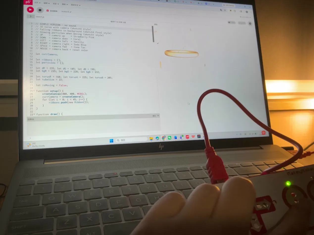 Hand holds Makey Makey while a pink torus and falling ribbons play on the screen with a mic button visible.