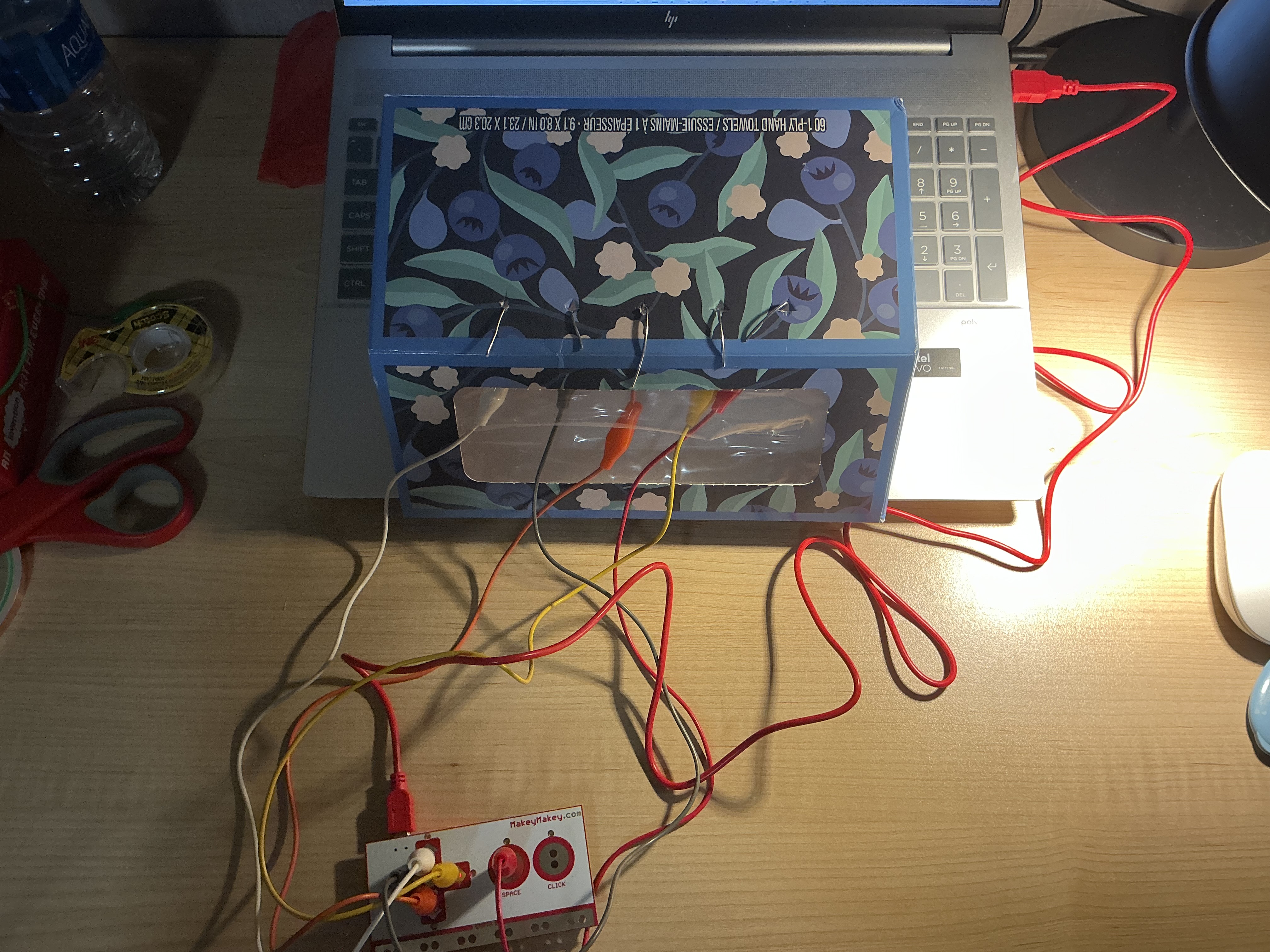 The image displays a top-down view of the blue floral box sitting on the laptop with the Makey Makey positioned below it and wires connecting both, showing the integrated physical setup from above.