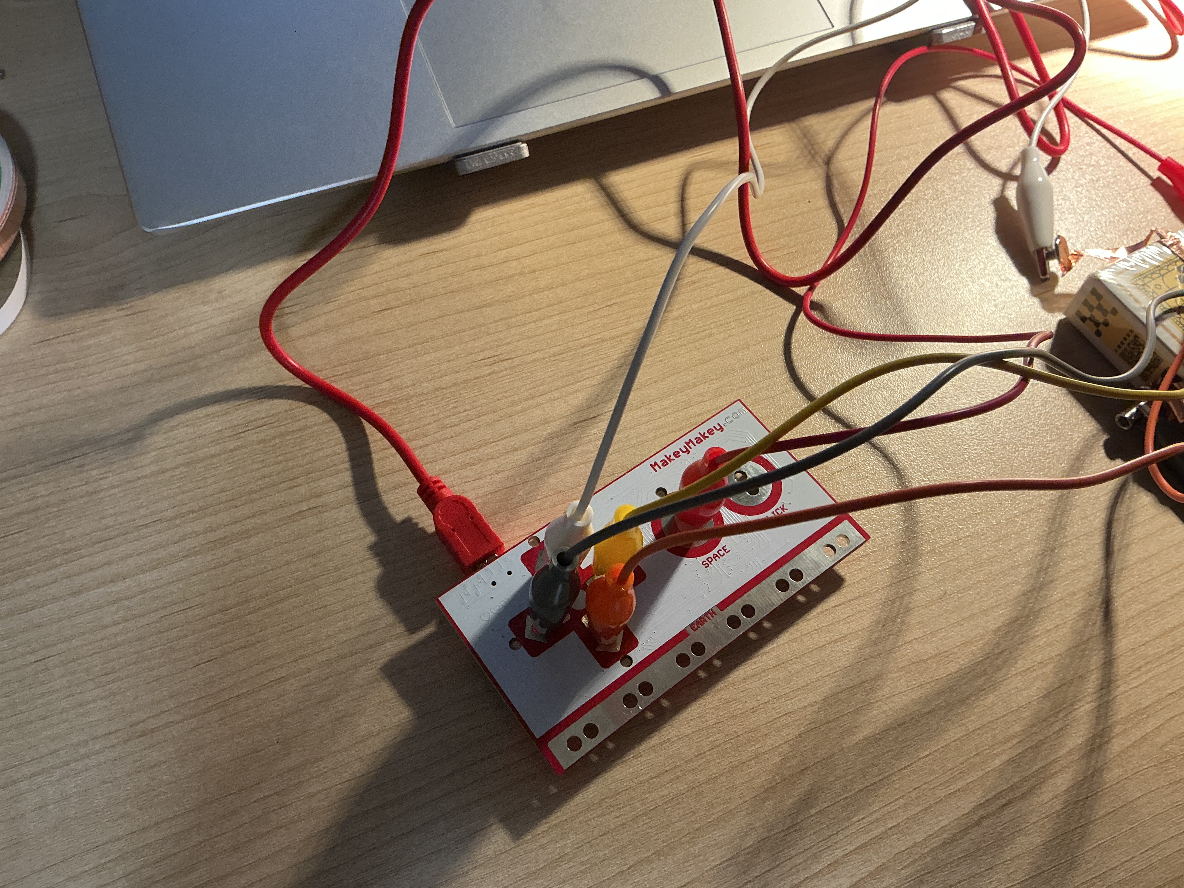 The image displays a top-down view of the Makey Makey board with multiple colored alligator clip wires spreading outward toward the yellow box interface, showing the full circuit wiring layout.