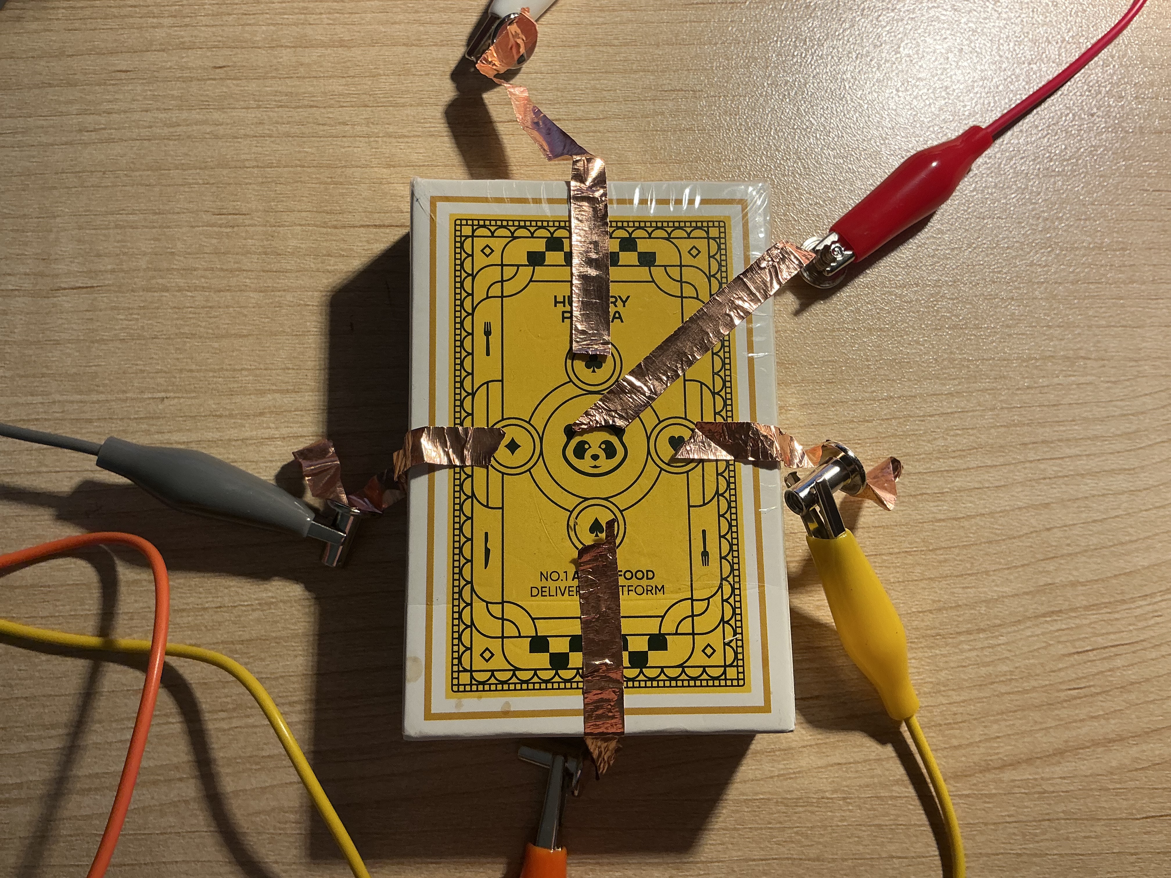 The image displays the yellow box viewed from above with the copper tape cross pattern visible and alligator clips attached at the edges, using copper tape strips as the directional conductive trigger surface.