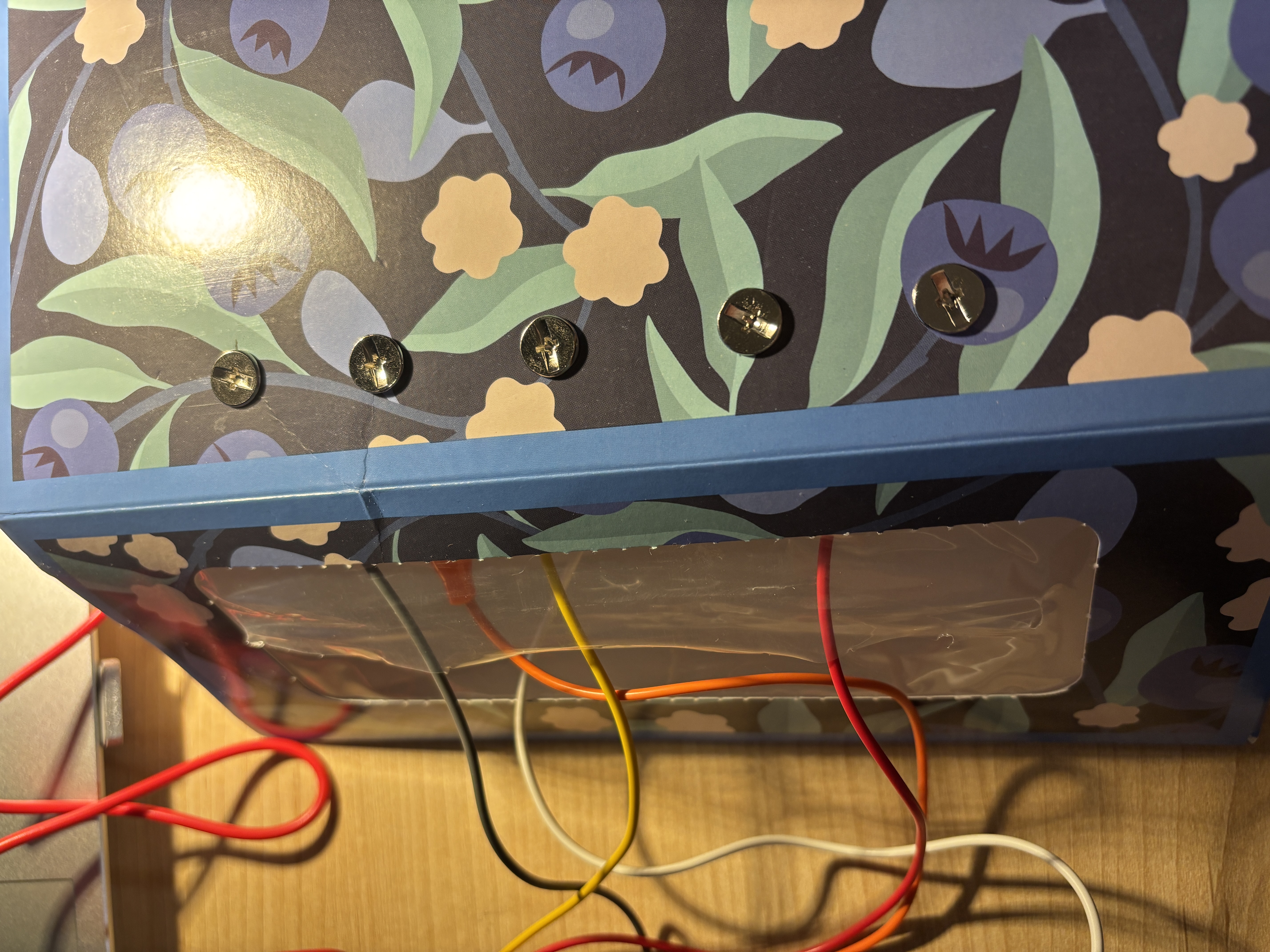 The image displays a close-up side view of the floral box top edge showing five iron wire loops pushed vertically through the surface, using bent iron wire as the raised conductive buttons.