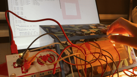 The image displays a hand pressing the iron wire loop buttons on the floral box while a visual response appears on the laptop screen, using iron wire and copper tape as the conductive trigger system.