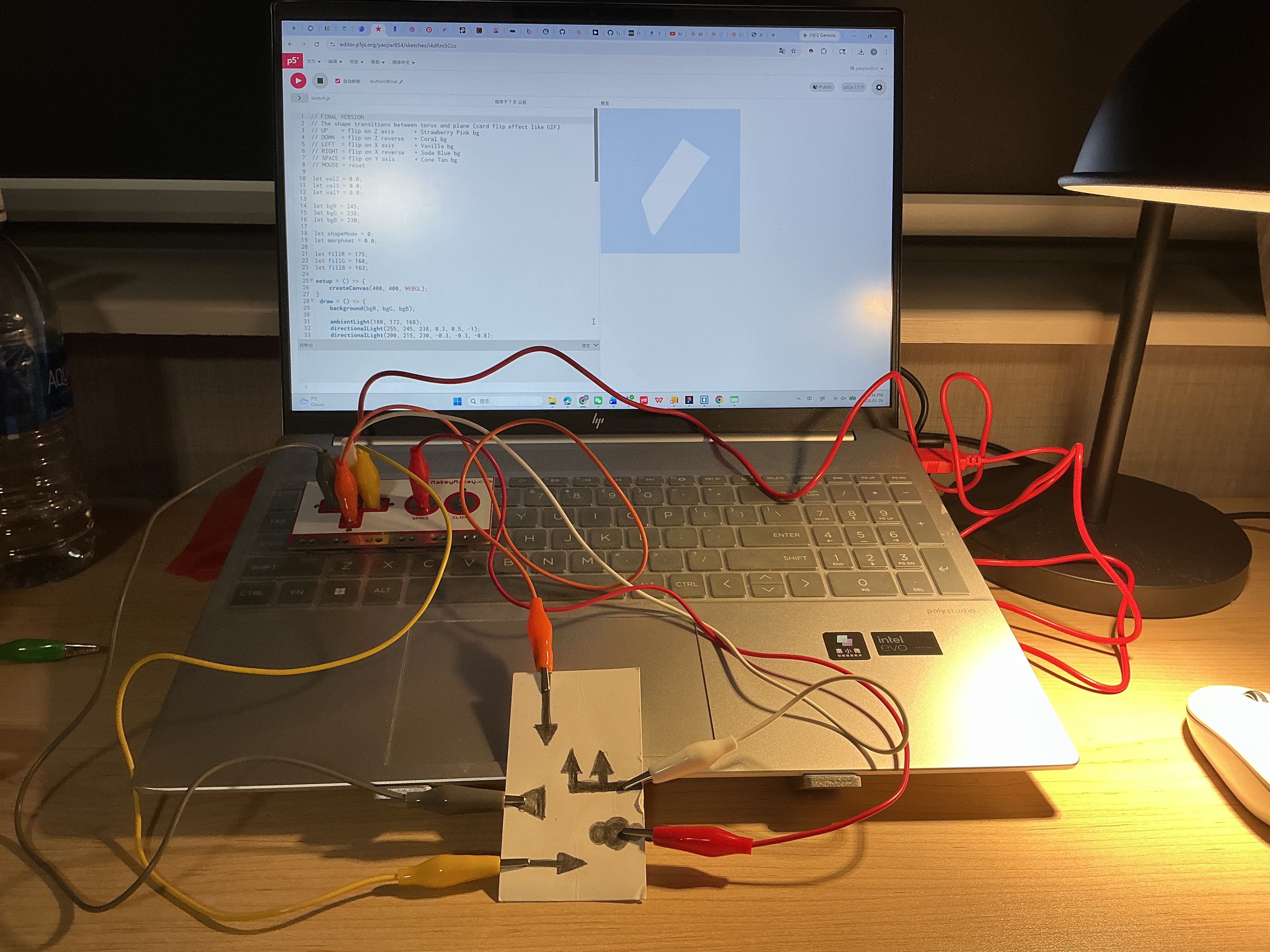 The image displays a laptop showing a white tilted flat plane rotating in 3D space with the pencil-drawn arrow card placed below, confirming graphite marks are successfully triggering the WEBGL sketch.