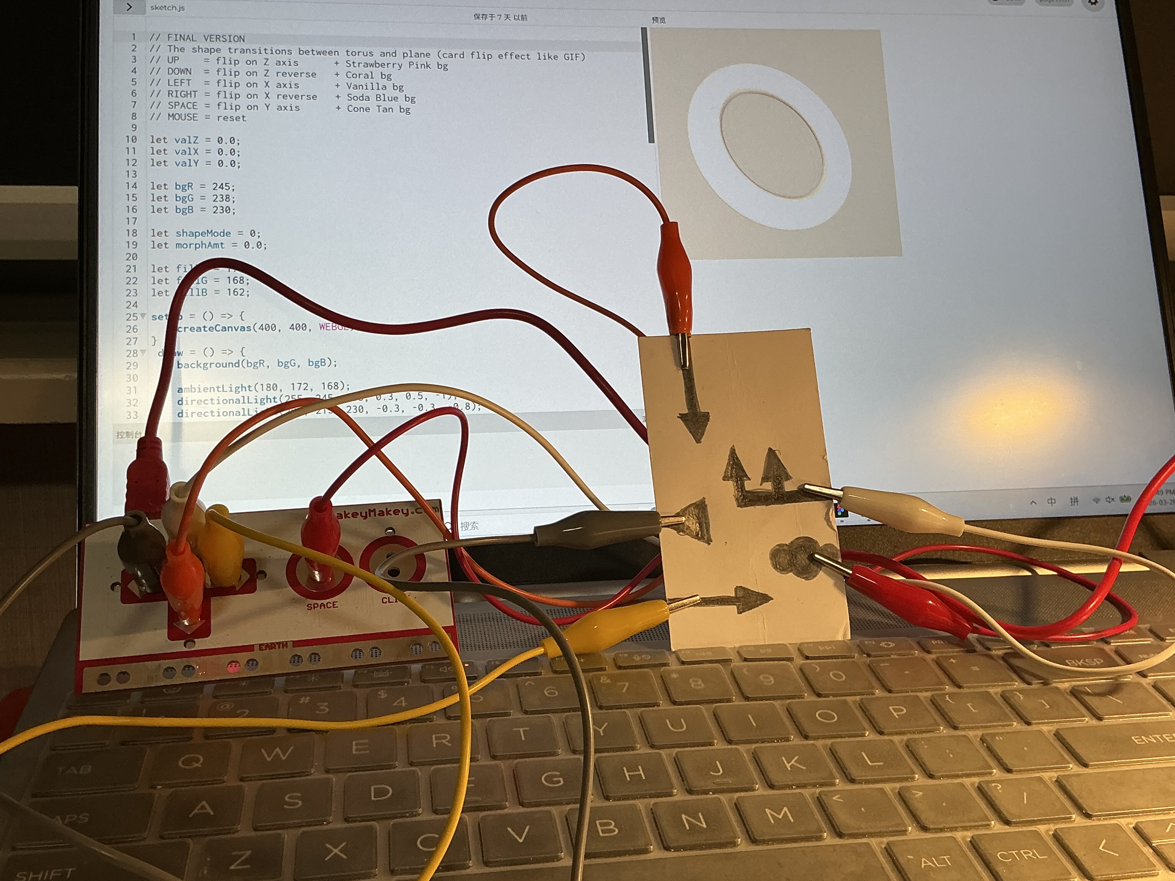 The image displays the pencil arrow card lying flat beside the Makey Makey board with a 3D torus visible on the laptop screen, using pencil graphite as the five-point directional conductive interface.